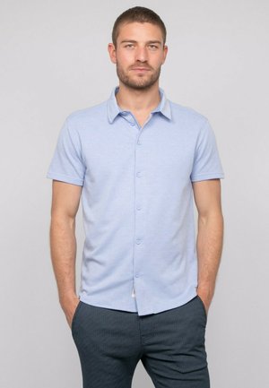 Homme aux cheveux courts et à la barbe, portant une chemise à manches courtes bleu clair boutonnée et un pantalon texturé foncé, mains dans les poches, fond neutre.