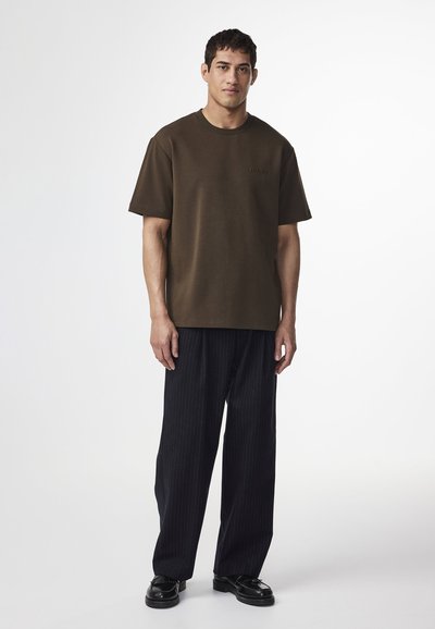 Oliven grønne bomulds t-shirt med rund hals, korte ærmer og et diskret logo; parret med sorte stribede bukser og sorte sko.