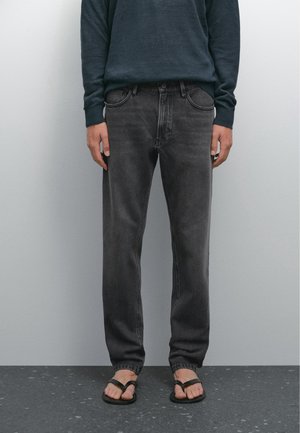 Henkilö, jolla on päällään tummanharmaat farkut, tumma pitkähiha-paita ja mustat varvassandaalit, seisoo vaaleanharjan tasaisen seinän edessä.