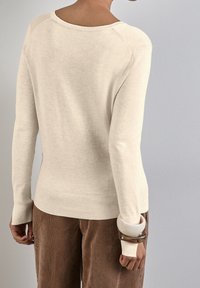 Un pull beige à manches longues avec un grand col et un ourlet côtelé, associé à un pantalon en velours côtelé marron. Un bracelet épais porté au poignet.