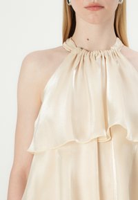 Beige halter top made of shiny satin, featuring a gathered neckline and layered ruffles that drape softly down the front.
