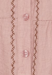 Lyserød stofskjorte med to klare knapper, lodret søm i brun og tekstureret materiale med en blød finish.