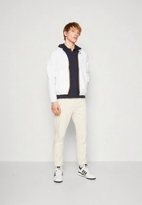 Hvit jakke med glidelås og hette, marineblå polotrøye, beige bukser og hvite joggesko med svarte detaljer. Enkel, uformell design.