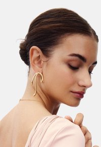 Guldmetallörhängen med en förlängd, böjd design. Slät yta, med en slingrande accent upptill. Modellen bär ett mjukt rosa tyg.
