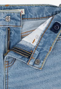 Jasnoniebieskie spodnie dżinsowe z zamkiem błyskawicznym metalowym, zapięciem na guzik oraz kontrastującym pomarańczowym przeszyciem. Widoczna podszewka na zewnątrz.