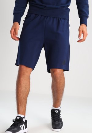 Man draagt marineblauwe Adidas-shorts, zwart-witte Adidas-sneakers en witte sokken, staand met ontspannen handen.