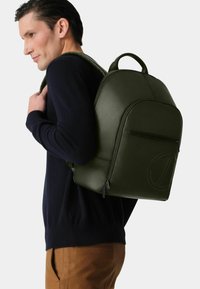 Ein Mann trägt einen dunklen Sweater und braune Hosen und hat einen großen olivgrünen Rucksack mit einer Vorderreißverschlusstasche und einem geprägten Logo.