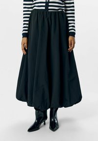 Jupe ballon noire, taille genoux, fabriquée en tissu texturé ; elle présente une ceinture élastique et est associée à des bottes noires à talons hauts et pointues.