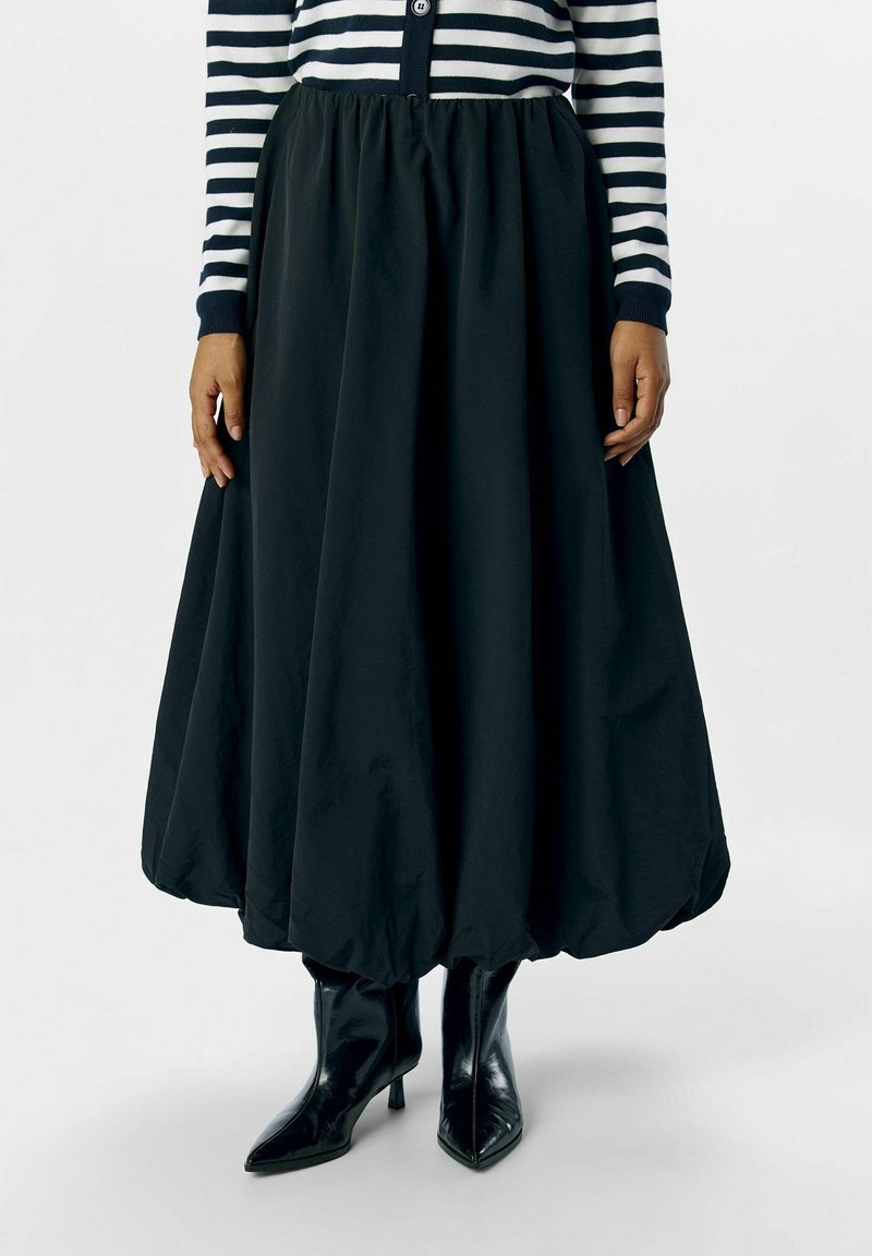 Jupe ballon noire, taille genoux, fabriquée en tissu texturé ; elle présente une ceinture élastique et est associée à des bottes noires à talons hauts et pointues.