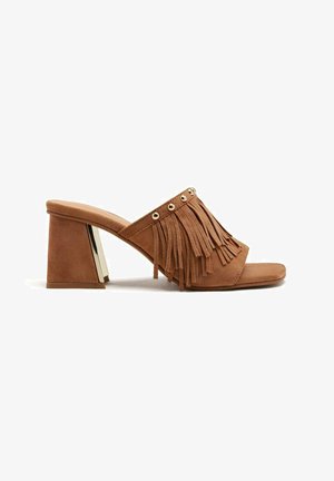 Mules à talons en daim beige avec un dessus à franges orné de clous dorés et un talon bloc avec un accent métallique. Design à bout ouvert.