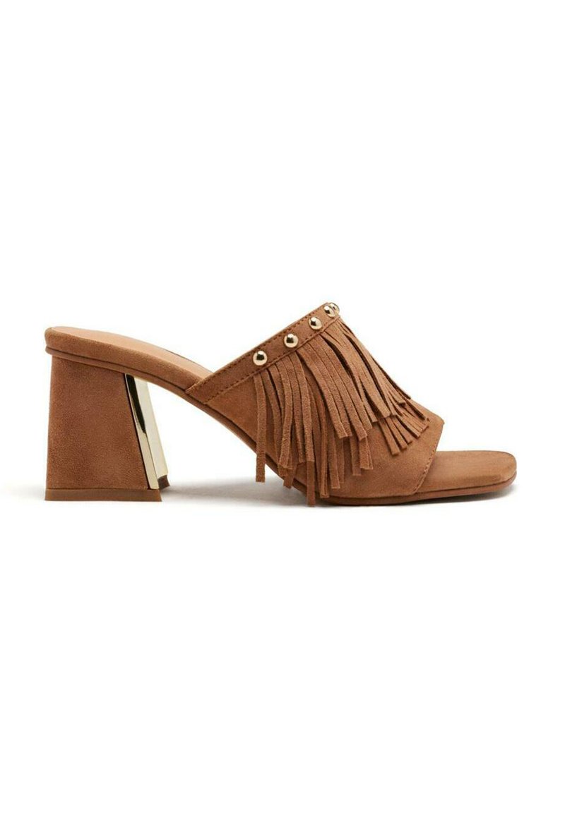 Mules à talons en daim beige avec un dessus à franges orné de clous dorés et un talon bloc avec un accent métallique. Design à bout ouvert.