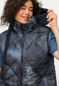 Gestepptes Weste in Dunkelblau mit metallic-glänzender Oberfläche, mit hohem Kragen, strukturiertem Stoff und Seitentaschen, über einem Denim-Hemd.