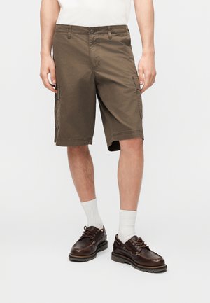Mann trägt braune Cargo-Shorts, weiße Socken und dunkelbraune Lederschuhe, steht vor einem schlichten Hintergrund.