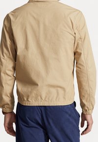 Krémová bunda s uvolněným střihem, vybavená elastickými manžetami a lemem, vyrobená z hladkého materiálu. Zadní strana má jednoduchý design bez ozdob.