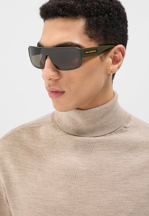 Giovane uomo che indossa occhiali da sole oversize verde oliva e dolcevita beige, di profilo su uno sfondo uniforme.