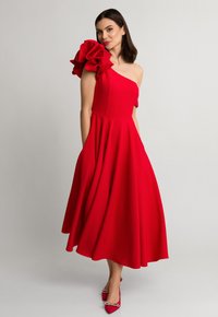 Czerwona sukienka na jedno ramię z dużym falbaniastym rękawem. Sięga poniżej kolan, z dopasowanym gorsetem i rozkloszowanym dołem. Posiada boczne kieszenie.
