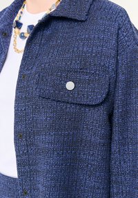 Giacca blu texturizzata con colletto, tasca a pattina con bottone e bottoni a pressione. Il tessuto presenta un motivo intrecciato per un ulteriore profondità.