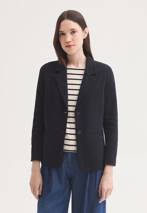 Femme portant un blazer noir par-dessus une chemise blanche à rayures noires et un jean bleu ample, debout devant un fond clair uni.
