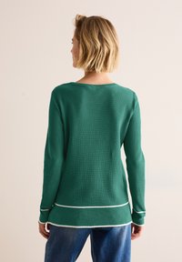 Grüner gerippter Pullover mit langen Ärmeln, der einen weißen unteren Streifen und einen Rundhalsausschnitt aufweist. Die Stofftextur wirkt weich und elastisch.
