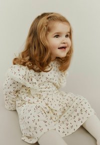 Kremasta haljina s malim crvenim cvjetnim uzorkom, nabranim rukavima s čipkastim rubom, mekana tkanina, uparena s bijelim tajicama. Dijete sjedi na podu.