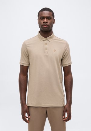 Joven hombre negro que lleva una camiseta tipo polo beige de manga corta y pantalones a juego, de pie frente a un fondo claro y liso.