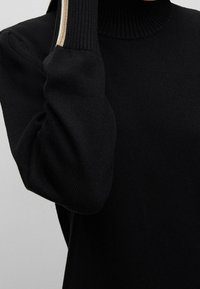 Czarny sweter robiony na drutach z wysokim, żebrowanym kołnierzem oraz strukturalnymi, objętościowymi rękawami. Posiada subtelny metaliczny akcent wzdłuż mankietu.
