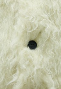 Krämfärgad ulltyg med en mjuk, fluffig textur, som har en rund svart knapp som en utmärkande detalj.