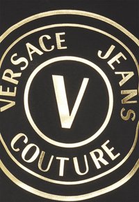 Musta kangas, jossa on ympyrämuotoilu ja kultateksti: "VERSACE JEANS COUTURE" sekä keskellä "V" -logo. Kultaiset yksityiskohdat ovat kiiltäviä ja heijastavia.
