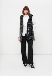 Zwarte puffer vest met een glanzende afwerking, voorzien van een capuchon en een verticale quiltstijl. Draag het over een wit shirt en combineer het met zwarte broek.