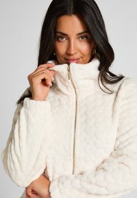 Kremowa polarowa kurtka z wysokim kołnierzem, teksturowanym wzorem w bąbelki i zamkiem z przodu. Miękki, pluszowy materiał z luźnymi rękawami.