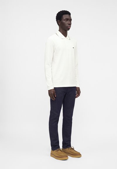Homme debout portant un polo blanc à manches longues, un pantalon foncé et des chaussures marron à lacets sur un fond clair uni.