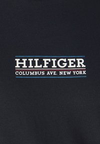 Tessuto nero con la scritta "HILFIGER" in bianco, con "COLUMBUS AVE. NEW YORK" in un font più piccolo sotto. Linee di accento blu e rosse.