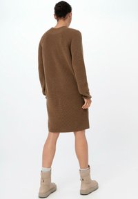 Dlouhé hnědé pletené šaty s ribbovanou texturou, kulatým výstřihem a dlouhými rukávy, končícími nad kolenem; nošeny s béžovými kotníkovými botami.