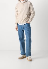 Beige huvtröja med frontficka över en vit skjorta med krage, matchad med ljusblå vida jeans och beige sportskor.
