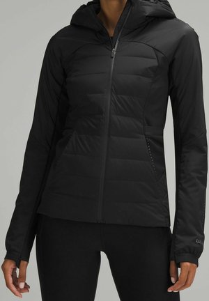 Femme portant une veste noire cintrée à capuche avec devant matelassé, fermeture zippée et passages pour le pouce aux manches, sur un fond gris uni.