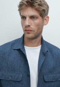 Uomo biondo con leggera barbetta guarda leggermente a sinistra, indossando una camicia blu con bottoni sopra una maglietta bianca, su uno sfondo chiaro e semplice.