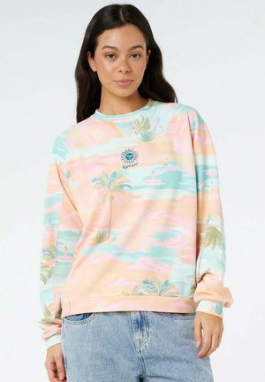 Frau mit langen dunklen Haaren, die einen pastellfarbenen Pullover mit tropischem Muster und hellblaue Jeans trägt, steht vor weißem Hintergrund.