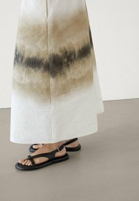 Sorte flade sandaler med en stropdesign, parret med en lang, flagrende, cremefarvet kjole med et gradientmønster i brun og sort.