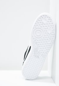 Schwarzer und weißer Sneaker mit einer strukturierten weißen Gummisohle, die kreisförmige Muster und das Puma-Logo an der Seite zeigt.