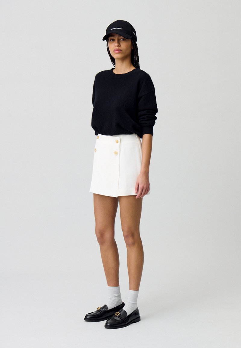 Suéter negro, falda corta blanca con botones dorados, mocasines negros y calcetines grises. El modelo lleva una gorra negra y se encuentra frente a un fondo neutro.