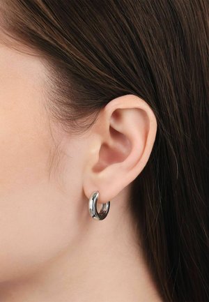 Nahaufnahme des linken Ohrs einer Person mit glattem braunem Haar, die einen kleinen, glänzenden silbernen Creolenohrring am Ohrläppchen trägt.