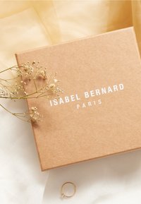 Brauner Kartonbehälter mit "ISABEL BERNARD PARIS" in weißem Text, neben einem goldenen Ring und getrockneten Blumen auf einem sanften weißen und gelben Hintergrund.
