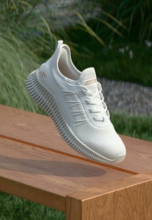 Chaussure de sport blanche Skechers avec semelle texturée et tige en mesh, suspendue au-dessus d'un banc en bois en plein air avec un fond d'herbe verte.