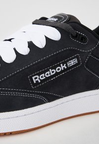 Sapatilhas de camurça preta com atacadores brancos, apresentando um logótipo da Reebok, costuras em contraste e uma sola de borracha branca texturizada com um destaque castanho.