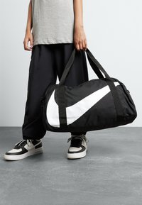 Black duffel bag with a white Nike swoosh, featuring sturdy fabric, dual handles, and a zippered closure, resting on a gray surface.