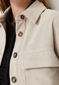 Beige corduroy jacket with a collared neck, featuring large brown buttons, a chest pocket, and ribbed texture.