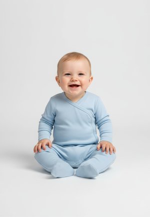 Hymyilevä vauva istuu valkoisella alustalla, pukeutuneena vaaleansiniseen, jalkoihin ulottuvaan potkupukuun, katse eteenpäin, kädet lepäävät polvilla.