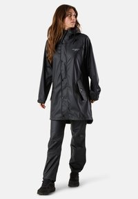 Schwarze wasserdichte Regenjacke mit Kapuze, ausgestattet mit einem Reißverschluss und Druckknöpfen, kombiniert mit passenden wasserdichten Hosen und schwarzer Fußbekleidung.