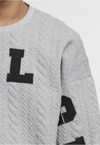 Pulover gri texturat, cu modele diagonale matlasate, având inscripții mari cu litere negre "L" pe piept și un "O" parțial vizibil pe mânecă.