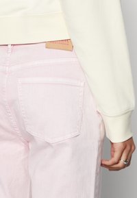 Calças jeans rosa claro com um corte de altura média, apresentando bolsos traseiros e uma etiqueta de couro na cintura. Textura de denim suave.